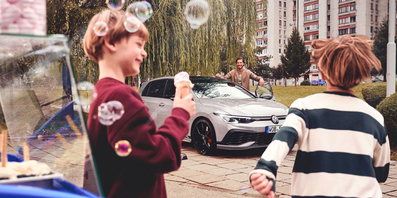Zwei Kinder stehen nahe einem Eisstand und halten Eis in den Händen, während sie auf einen geparkten silbernen Volkswagen Golf blicken. Im Hintergrund ist eine dritte Person, die am Auto steht. 