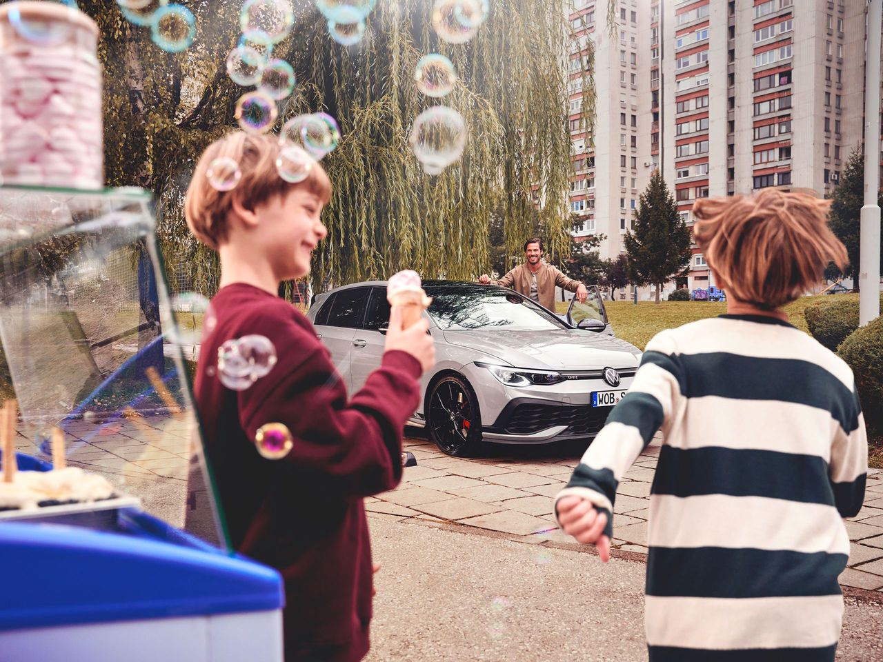 Zwei Kinder stehen nahe einem Eisstand und halten Eis in den Händen, während sie auf einen geparkten silbernen Volkswagen Golf blicken. Im Hintergrund ist eine dritte Person, die am Auto steht. 