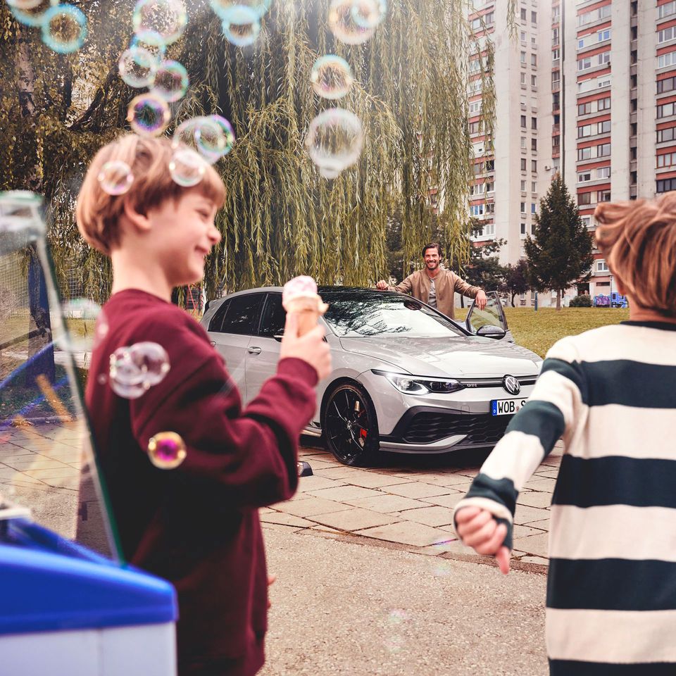 Zwei Kinder stehen nahe einem Eisstand und halten Eis in den Händen, während sie auf einen geparkten silbernen Volkswagen Golf blicken. Im Hintergrund ist eine dritte Person, die am Auto steht. 