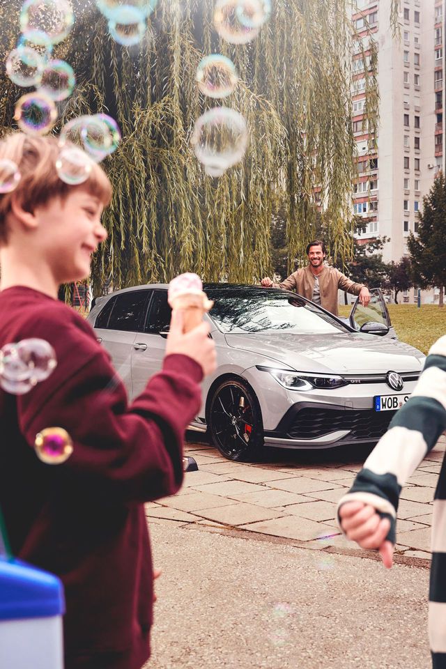 Zwei Kinder stehen nahe einem Eisstand und halten Eis in den Händen, während sie auf einen geparkten silbernen Volkswagen Golf blicken. Im Hintergrund ist eine dritte Person, die am Auto steht. 