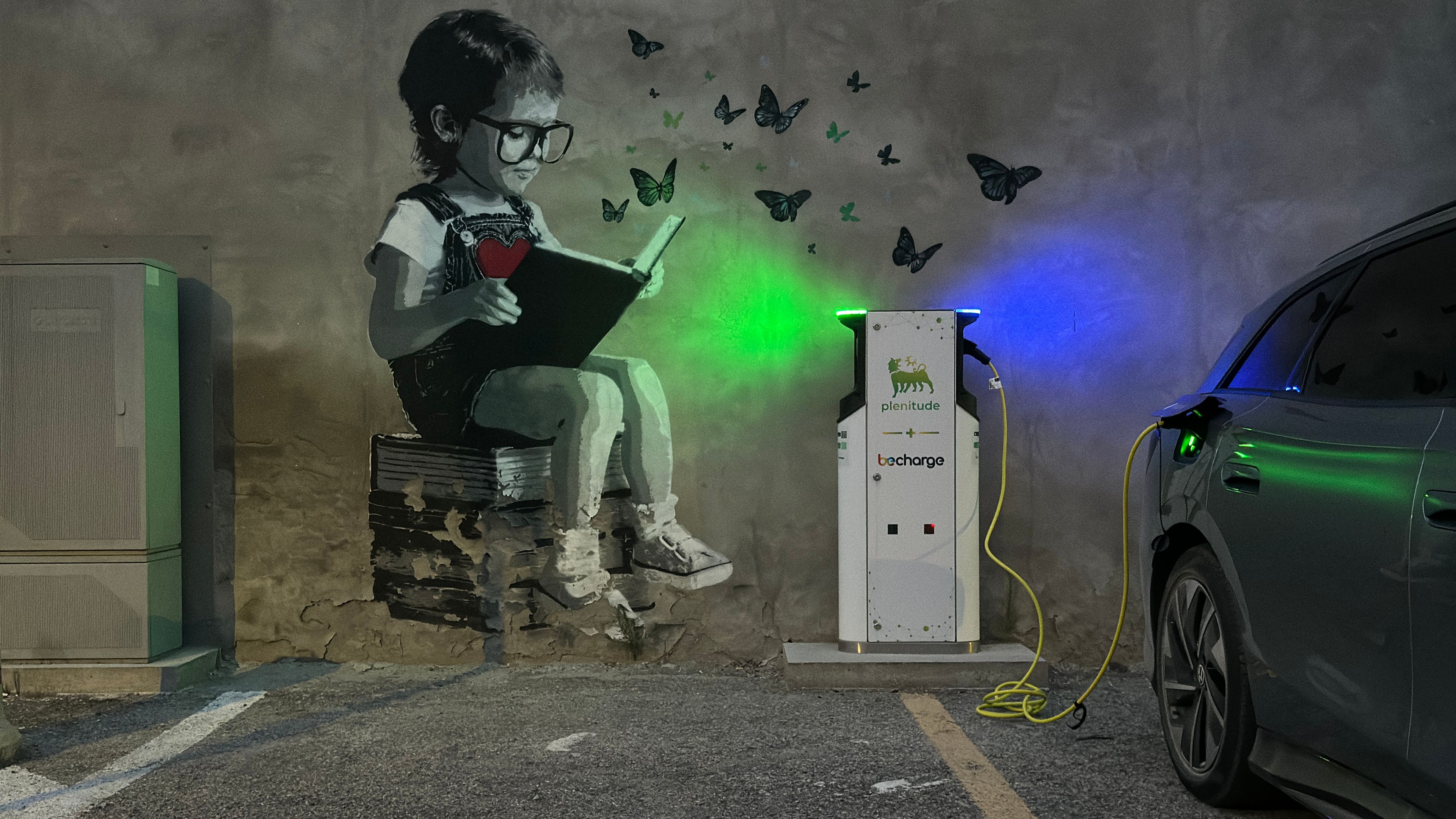 Ein Streetart-Gemälde zeigt ein Kind, das auf einem Stapel Bücher sitzt und beim Lesen Schmetterlinge freilässt. Daneben steht eine Elektro-Ladesäule mit einem angeschlossenen Auto. Das Bild ist bei Dunkelheit aufgenommen und die Ladesäule ist beleuchtet.