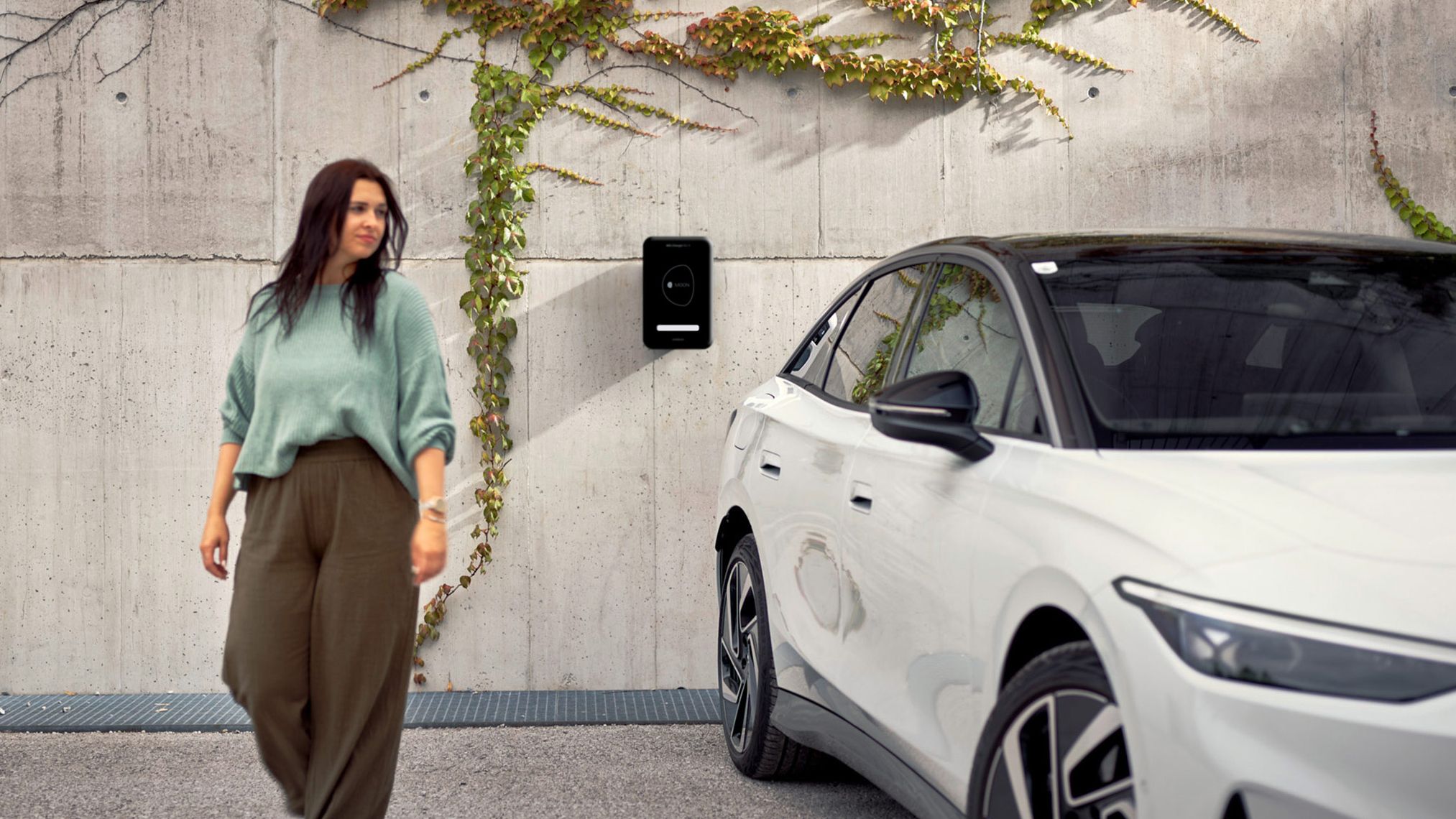 Eine Frau geht in der Nähe eines weißen ID.7, das an einer Wand mit einer Ladestation und Kletterpflanzen geparkt ist. Die Umgebung wirkt urban und modern.