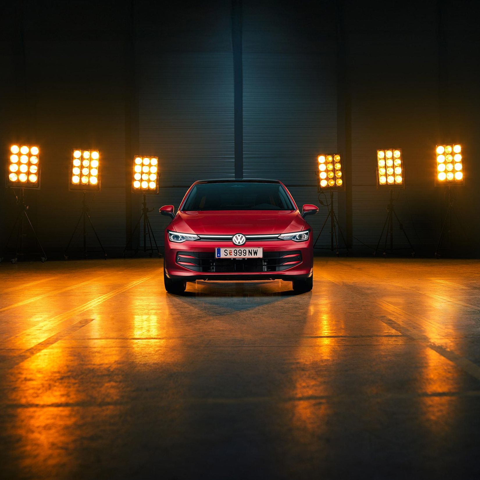 Ein roter VW Golf Falco Edition in Frontalansicht steht mittig im Bild in einer leeren Halle, Bühnenlichter strahlen das Fahrzeug von hinten an.