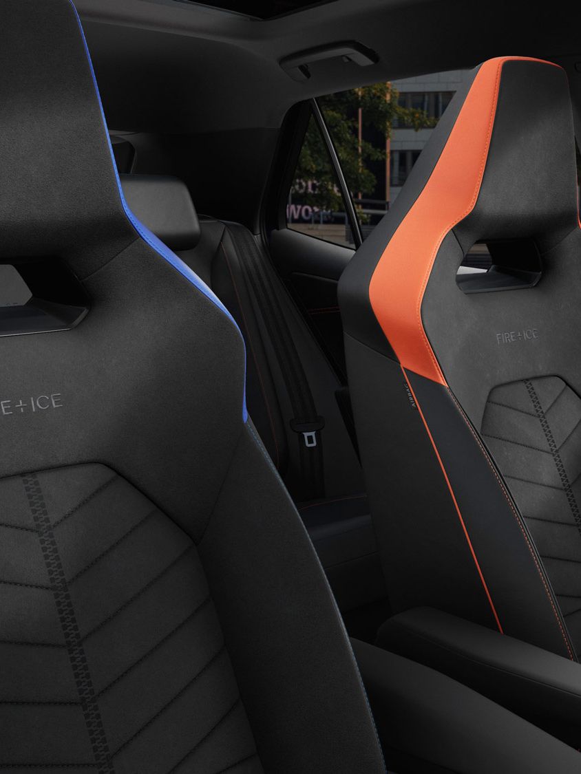 Zwei Rennsportsitze im ID.3 GTX FIRE+ICE, einer ist blau und der andere ist orange. Beide Sitze haben stilvolle Nähte und sind für Komfort und Unterstützung entworfen. Im Hintergrund sind Bäume und Gebäudestrukturen sichtbar.