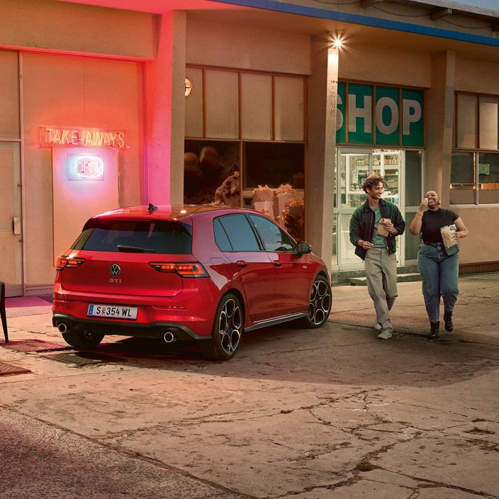 Roter VW Golf GTI steht abends vor einem Supermarkt, zwei junge Leute gehen mit Einkäufen daran vorbei.