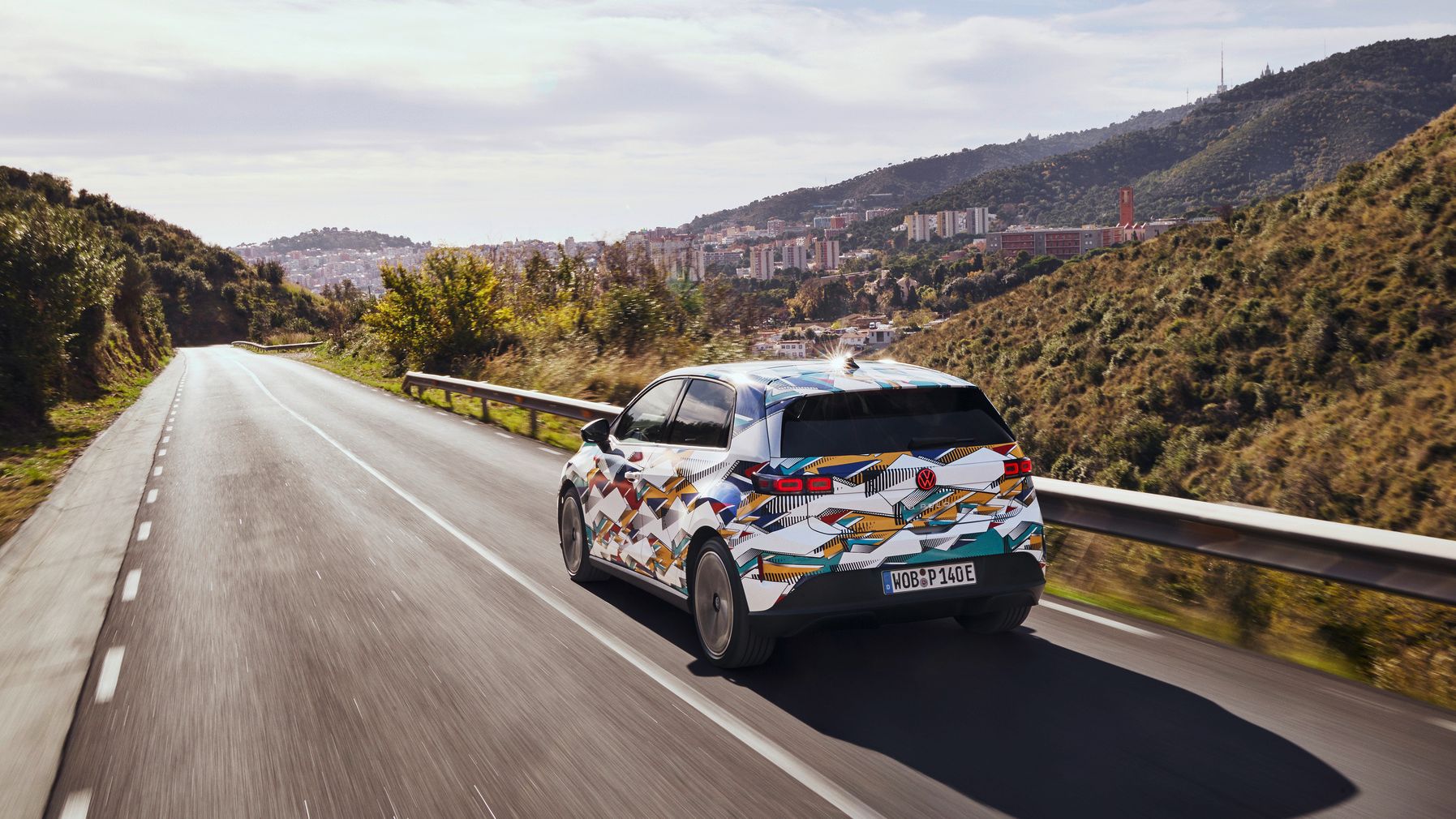 VW ID. Polo fährt auf einer Straße durch eine hügelige Landschaft mit Fernsicht auf eine Stadt. Die Straße ist von Bäumen und hügeligen Wiesen gesäumt. Der Himmel ist leicht bewölkt.