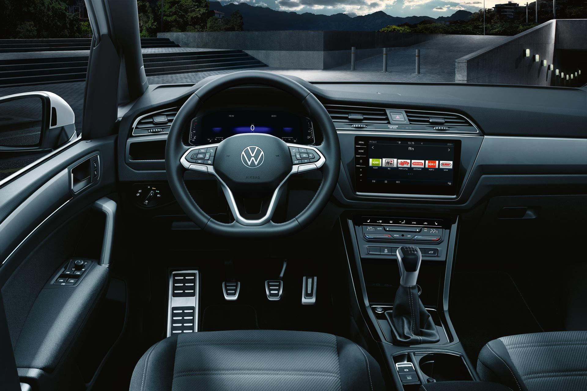 Innenraum des VW Touran mit schwarzem Leder. Das Bild zeigt das Lenkrad, die Mittelkonsole mit einem Touchscreen und einem manuellen Schalthebel. Der Wagen steht im Freien, vor einem Bergpanorama.
