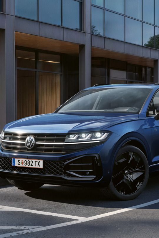 Ein blauer VW Touareg Final Edition steht vor einem Bürogebäude mit Glasfassade - Ansicht von schräg vorne.