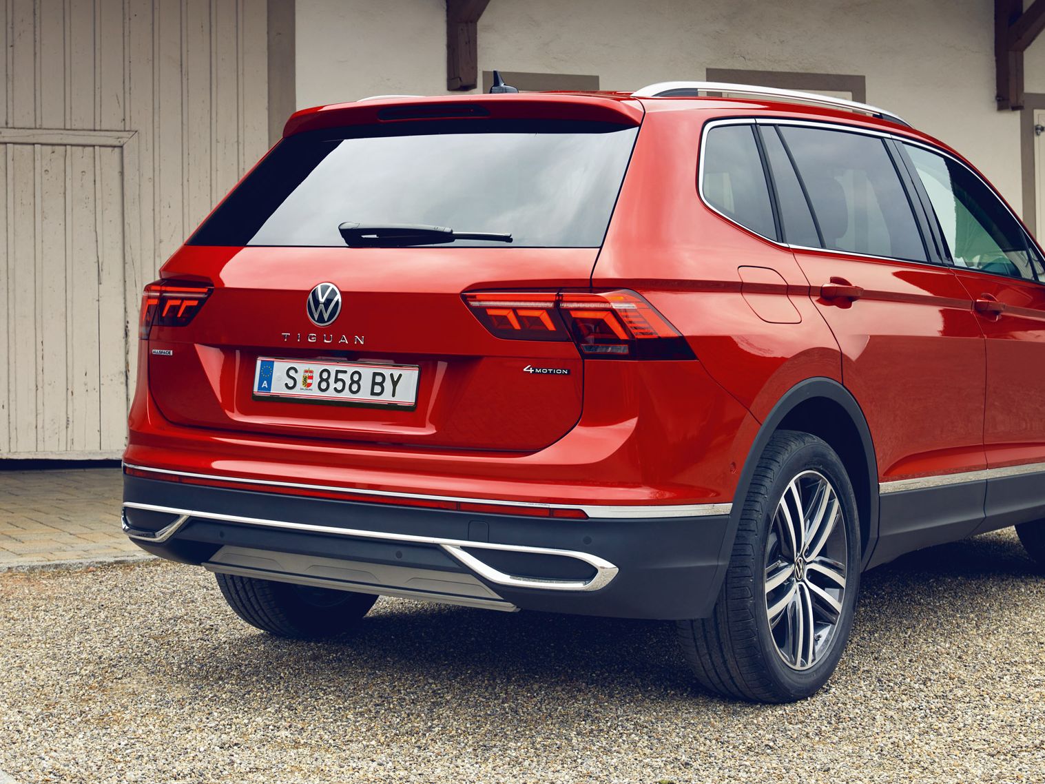 Roter VW Tiguan Allspace steht auf einem Kiesplatz vor einem hellen Gebäude mit Holztor. Das Auto ist aus schräger Heckansicht zu sehen, mit erkennbaren Rückleuchten, Modellschriftzug und österreichischem Kennzeichen.