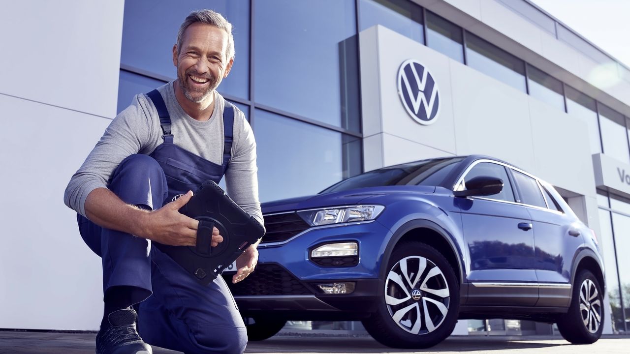 Ein Volkswagen Service Techniker mit Tablet, kniet vor einem blauen Volkswagen T-Roc