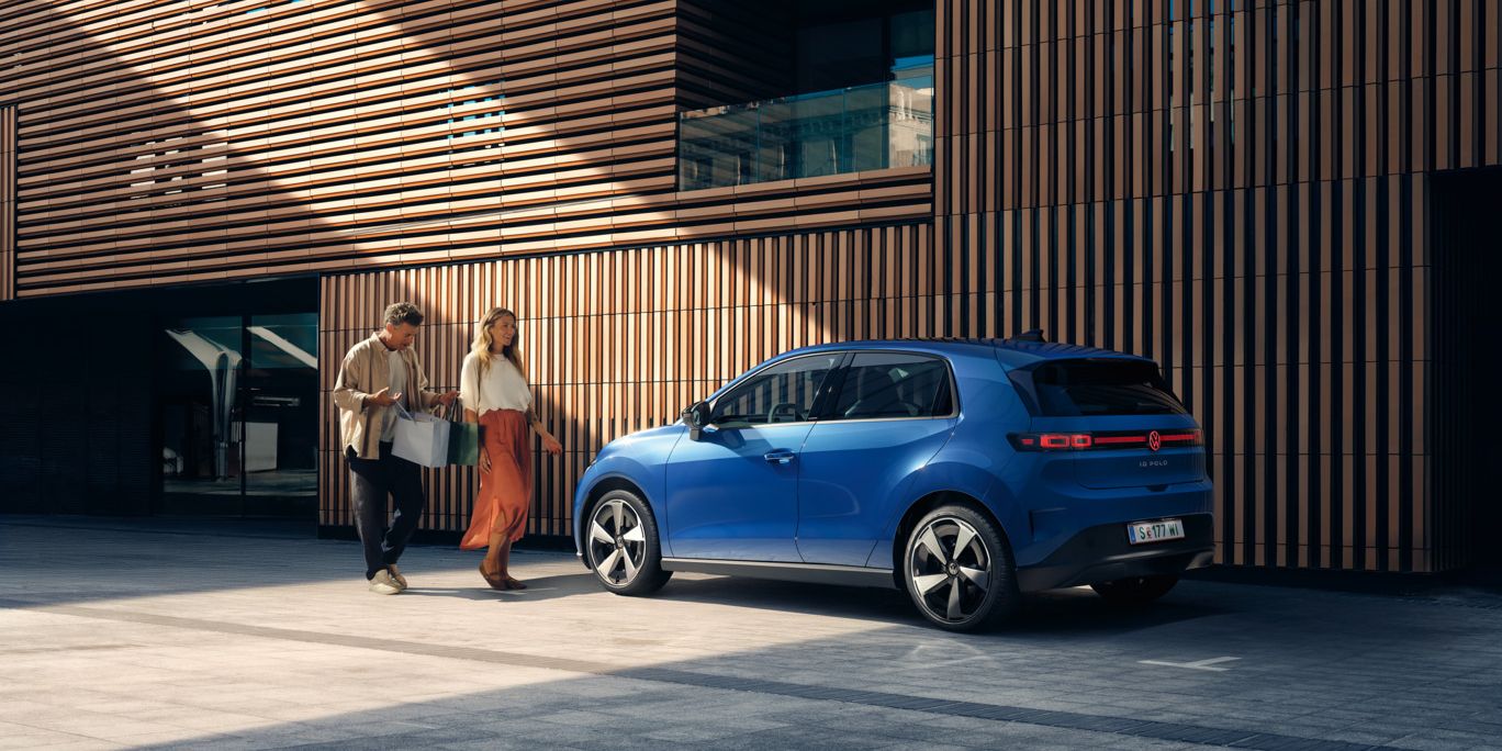 Ein blauer VW ID. Polo Style steht vor einem Gebäude mit Holzfassade. Ein Mann und eine Frau mit Einkaufstaschen gehen darauf zu