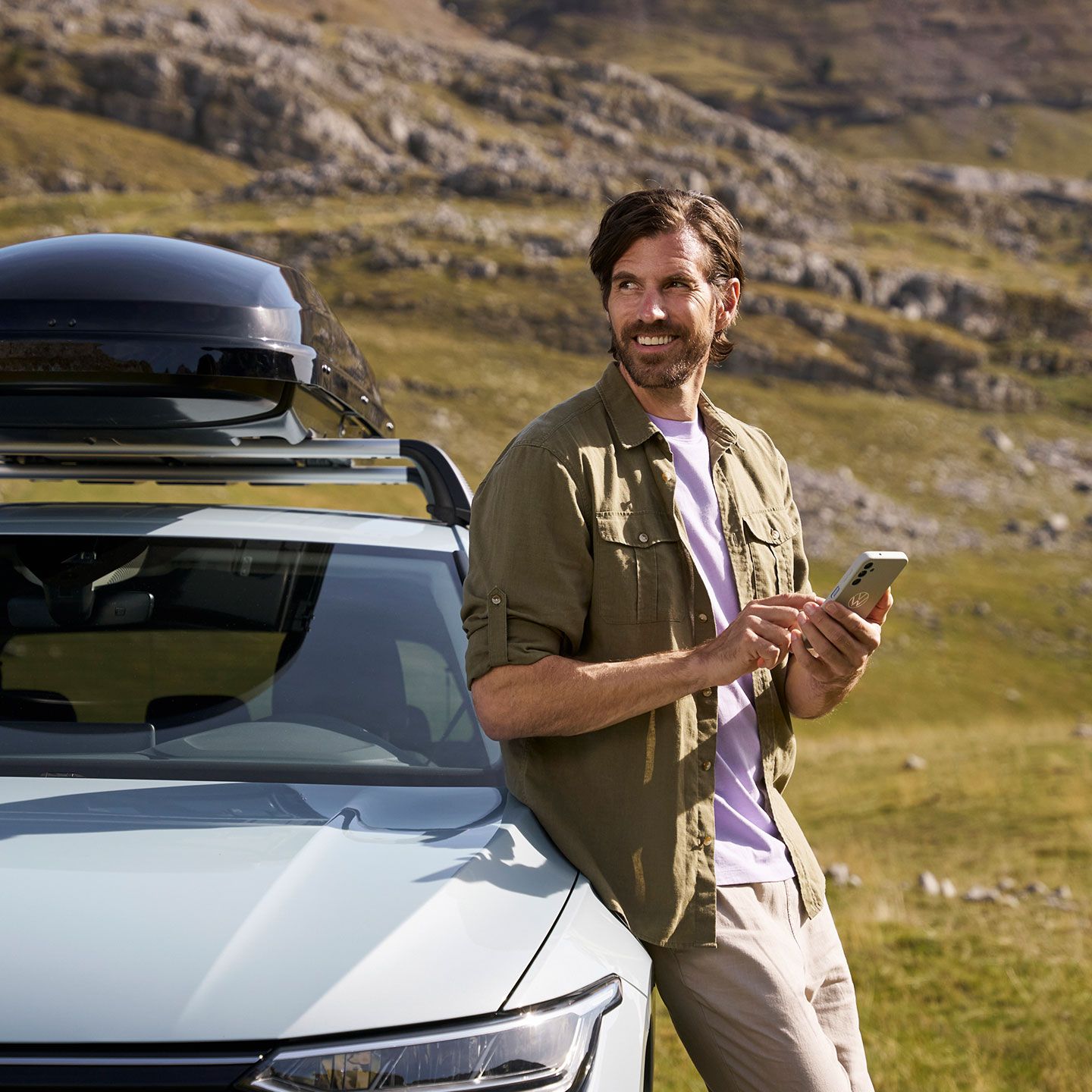 Ein Mann lehnt an einem silbernen VW Modell an und schaut in die Ferne, während er sein Handy in der Hand hält.