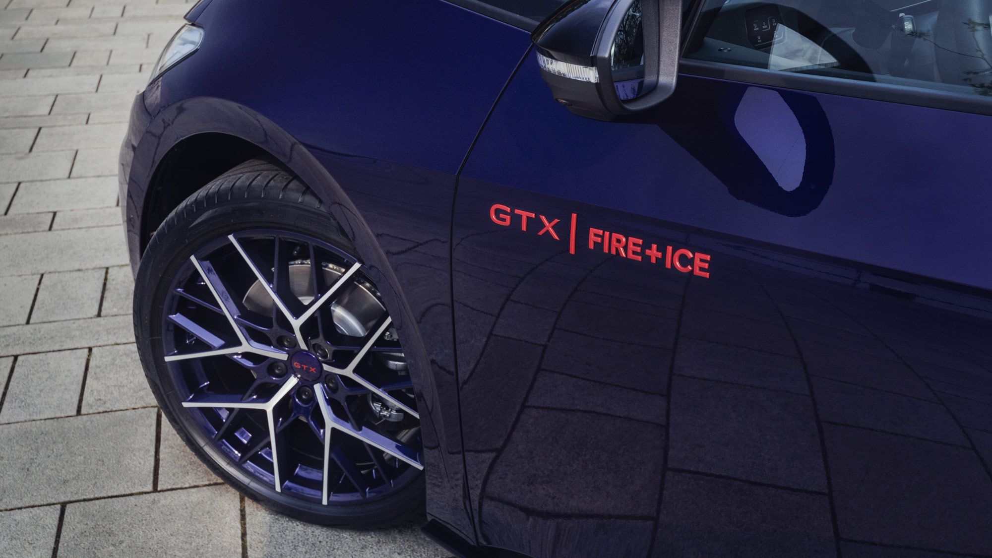 Nahaufnahme eines blauen Autos mit der Aufschrift "GTX FIRE+ICE" über dem Reifen. Die Felgen sind dunkel und das Auto steht auf einem gepflasterten Boden. Das Seitenspiegel sind teilweise sichtbar.