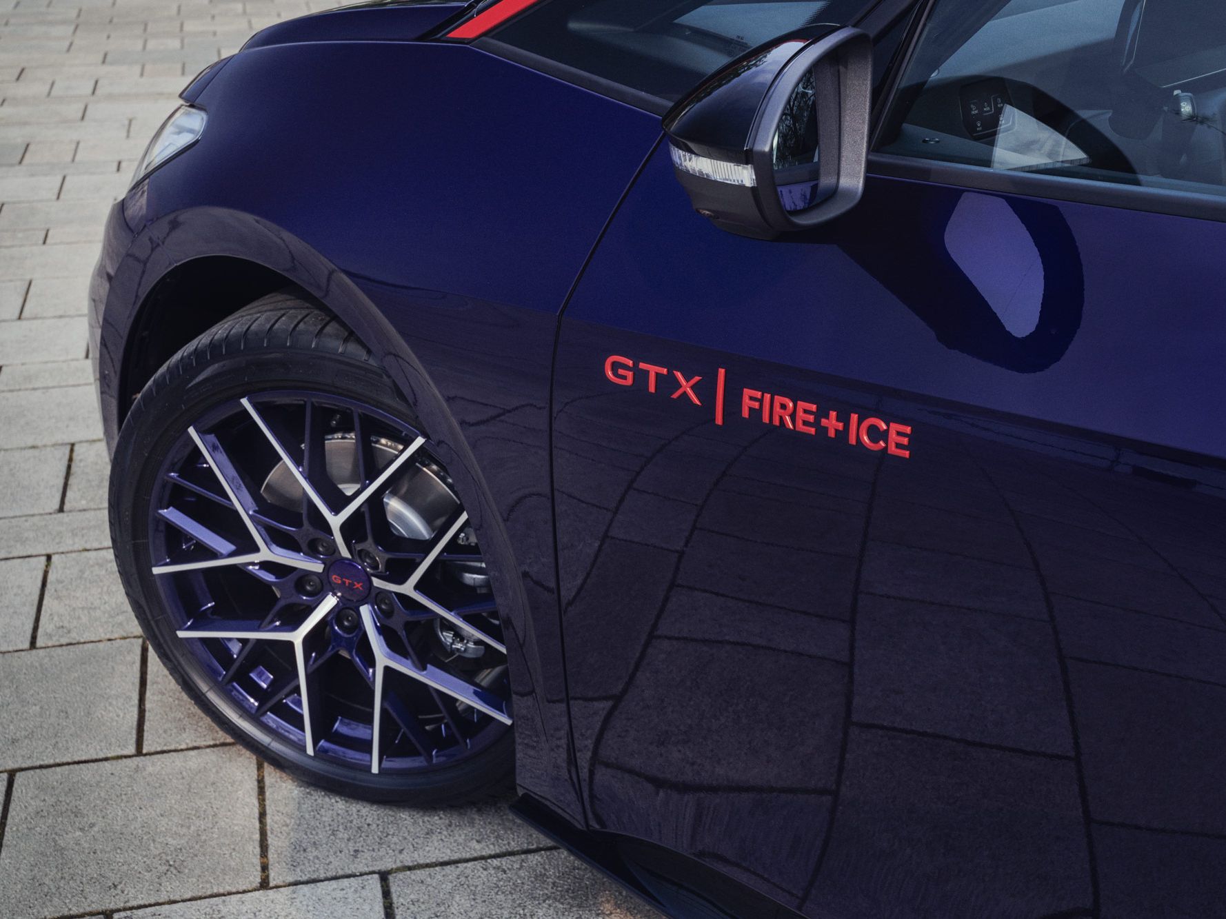 Nahaufnahme eines blauen Autos mit der Aufschrift "GTX FIRE+ICE" über dem Reifen. Die Felgen sind dunkel und das Auto steht auf einem gepflasterten Boden. Das Seitenspiegel sind teilweise sichtbar.