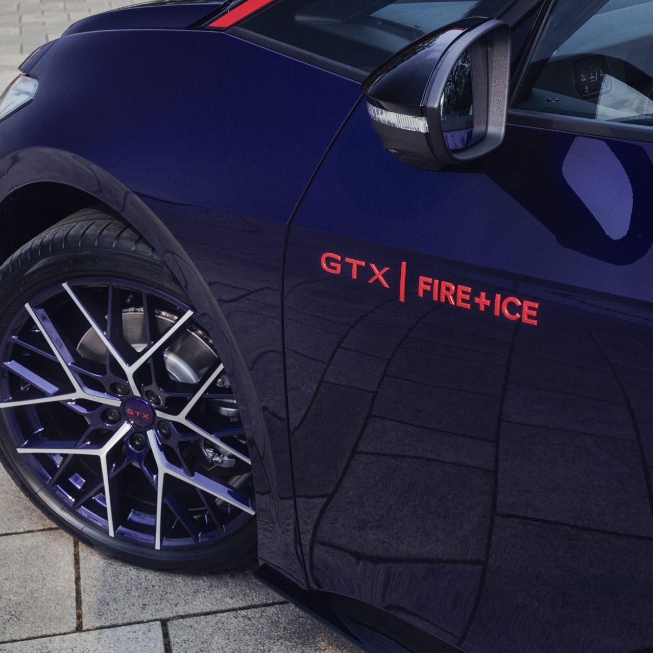 Nahaufnahme eines blauen Autos mit der Aufschrift "GTX FIRE+ICE" über dem Reifen. Die Felgen sind dunkel und das Auto steht auf einem gepflasterten Boden. Das Seitenspiegel sind teilweise sichtbar.