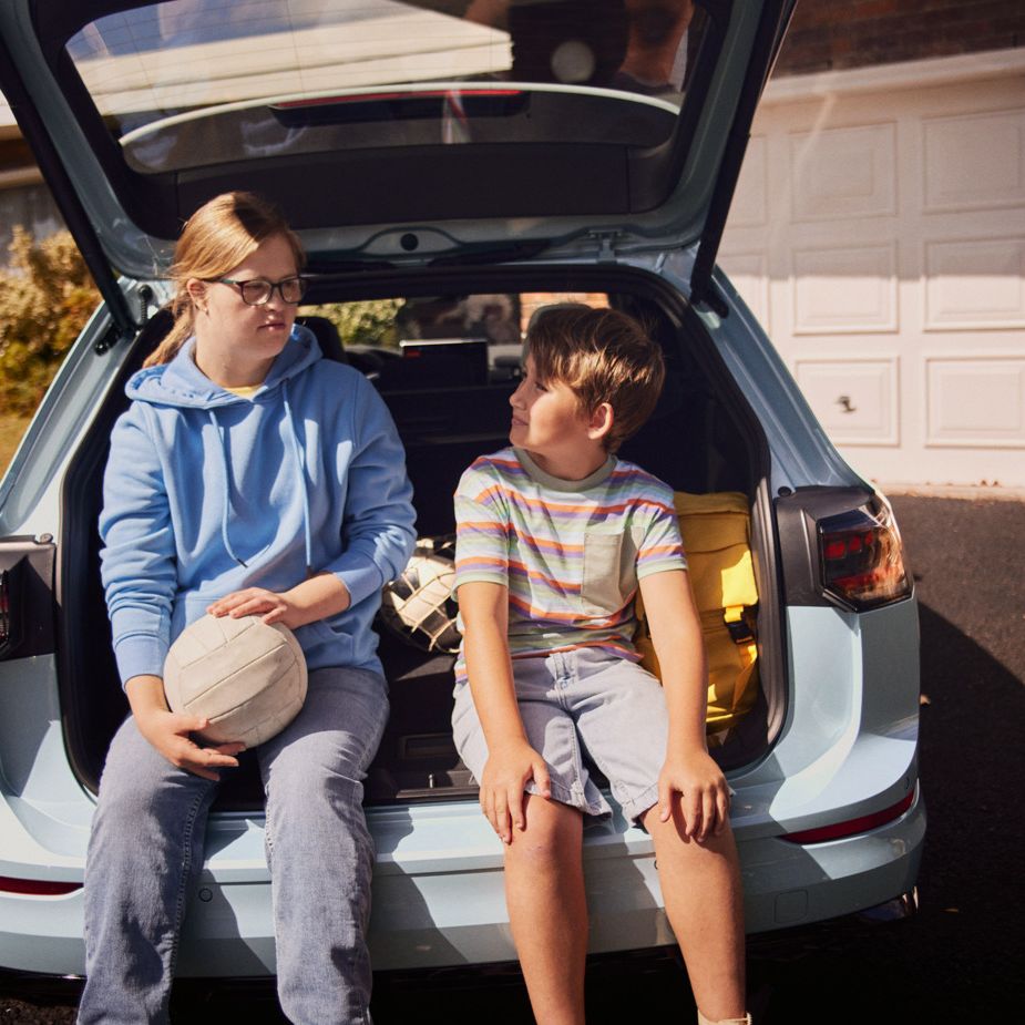 Junge und Mädchen sitzen im offenen Kofferraum eines VW Golf Variant in einer Auffahrt.