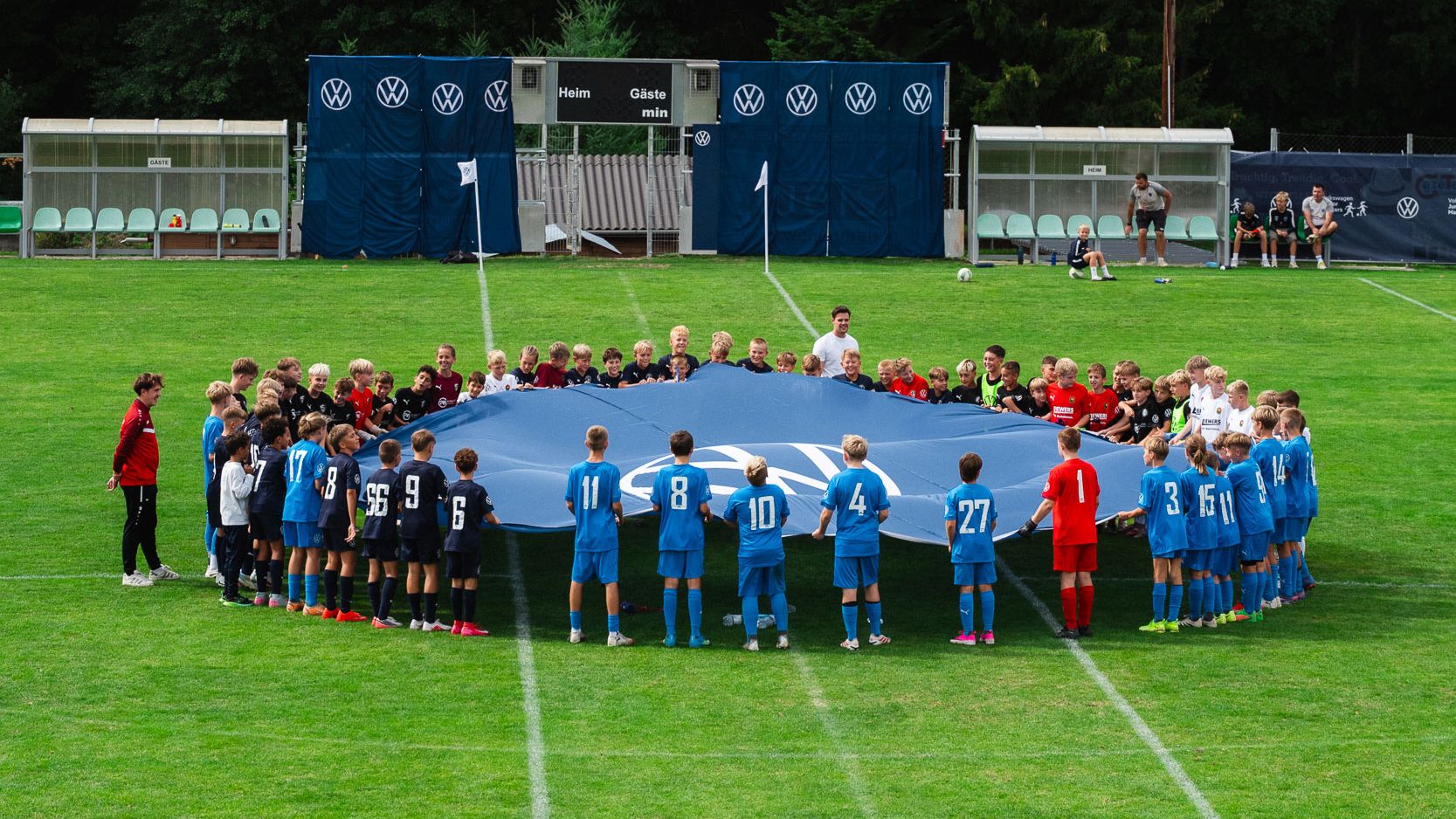 Eine große Gruppe von Kindern in verschiedenen Fußballtrikots steht im Kreis auf einem Fußballfeld und hält gemeinsam ein riesiges blaues Tuch oder Banner. Im Hintergrund sind Ersatzbänke, Werbebanner und Bäume zu sehen. Die Szene wirkt wie eine feierliche Eröffnung oder ein gemeinsames Teamritual vor einem Spiel.