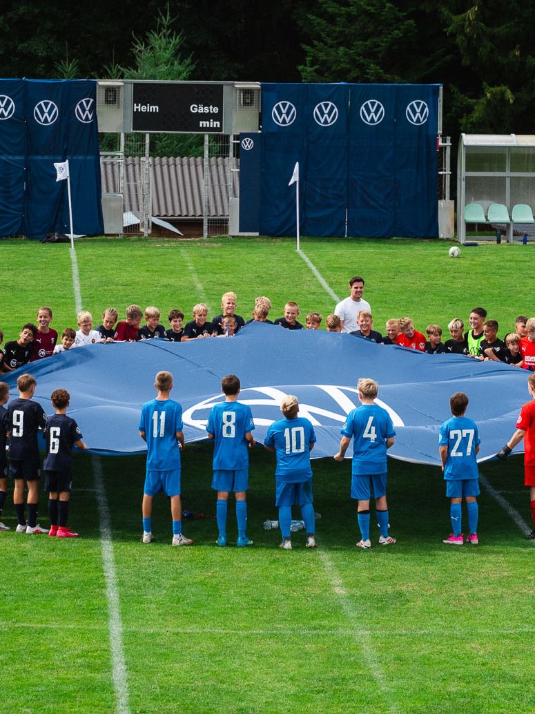 Eine große Gruppe von Kindern in verschiedenen Fußballtrikots steht im Kreis auf einem Fußballfeld und hält gemeinsam ein riesiges blaues Tuch oder Banner. Im Hintergrund sind Ersatzbänke, Werbebanner und Bäume zu sehen. Die Szene wirkt wie eine feierliche Eröffnung oder ein gemeinsames Teamritual vor einem Spiel.