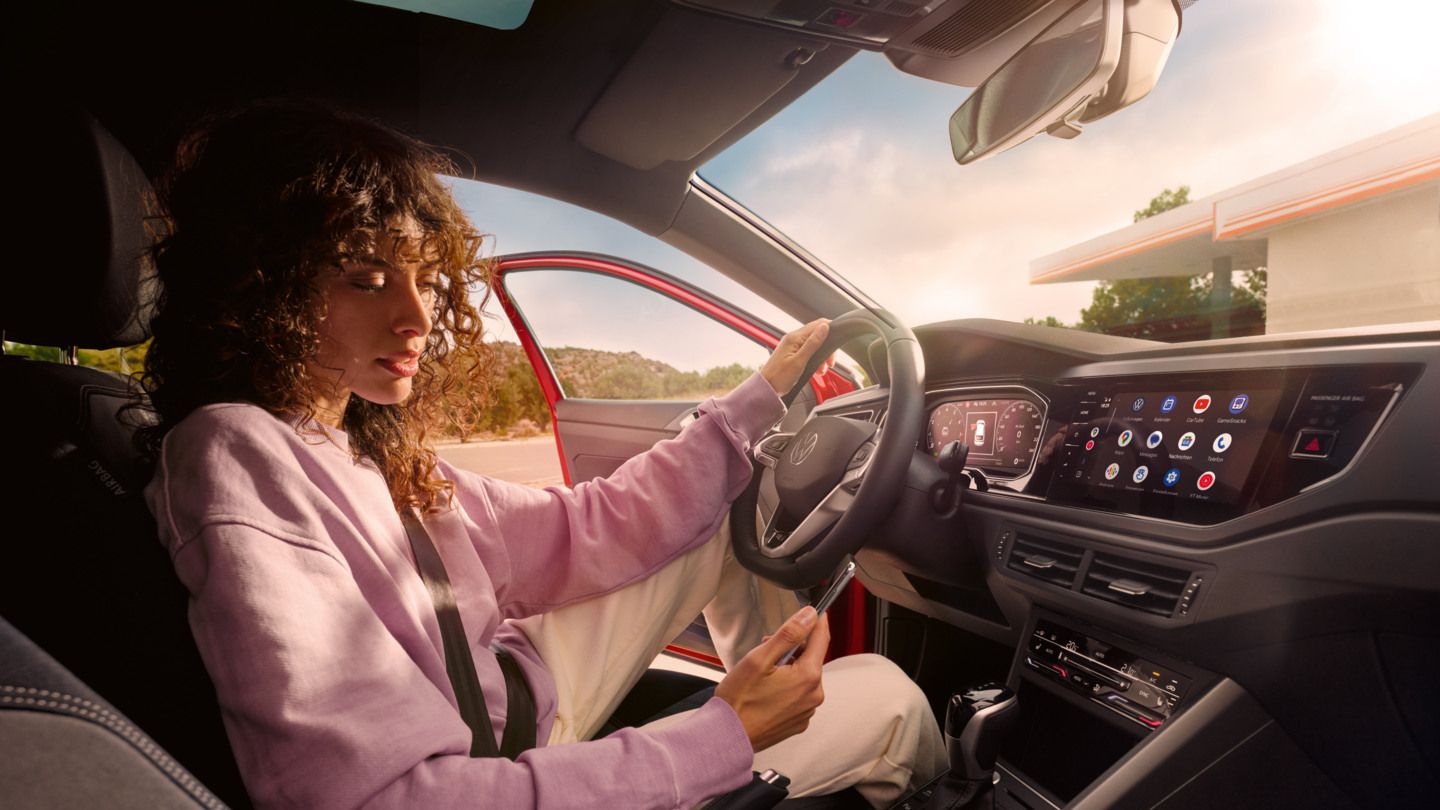 Person in einem VW Taigo sitzt auf dem Fahrersitz, trägt einen Sicherheitsgurt und hat eine Hand auf dem Lenkrad abgelegt, während sie mit der anderen ein Smartphone bedient. Auf der Mittelkonsole ist ein großes Display mit verschiedenen Apps und Funktionen zu sehen. Durch die geöffnete Fahrertür erkennt man im Hintergrund eine Tankstelle oder ein Rasthofgebäude.