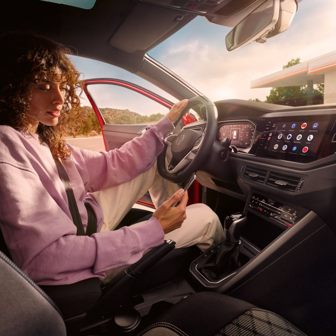 Person in einem VW Taigo sitzt auf dem Fahrersitz, trägt einen Sicherheitsgurt und hat eine Hand auf dem Lenkrad abgelegt, während sie mit der anderen ein Smartphone bedient. Auf der Mittelkonsole ist ein großes Display mit verschiedenen Apps und Funktionen zu sehen. Durch die geöffnete Fahrertür erkennt man im Hintergrund eine Tankstelle oder ein Rasthofgebäude.