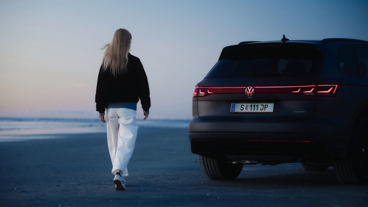 Eine Frau mit langen, blonden Haaren geht am Strand entlang bei Sonnenuntergang. Neben ihr steht ein schwarzer Volkswagen Touareg R mit leuchtenden Rücklichtern. 