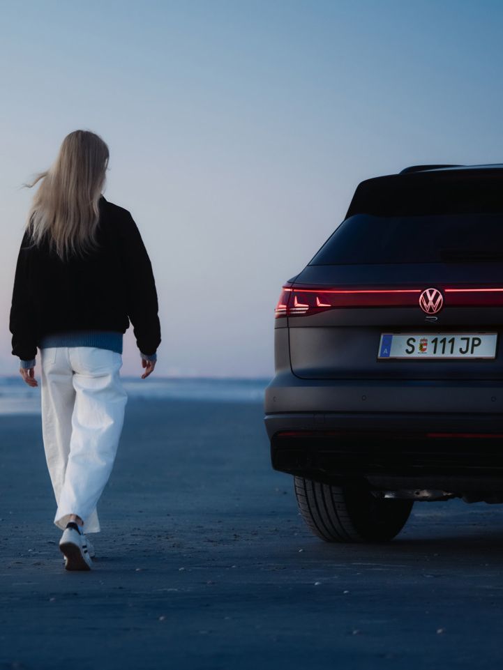 Eine Frau mit langen, blonden Haaren geht am Strand entlang bei Sonnenuntergang. Neben ihr steht ein schwarzer Volkswagen Touareg R mit leuchtenden Rücklichtern. 