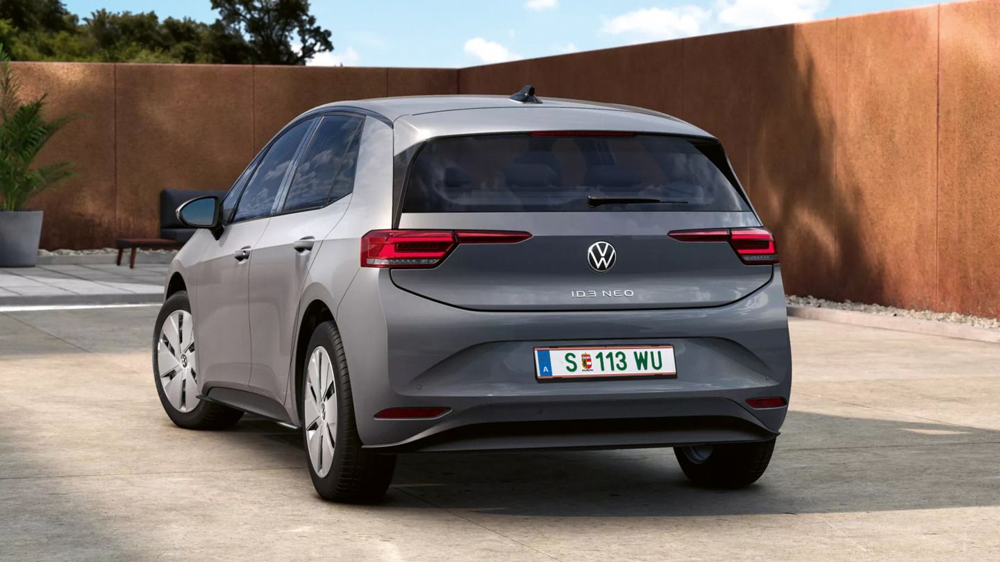 Ein grauer VW ID.3 Neo steht auf einem gepflasterten Hof, von hinten leicht seitlich aufgenommen. Die klaren LED-Rückleuchten und das VW-Logo sind gut sichtbar, das Auto wirkt modern und kompakt. Im Hintergrund sind eine hohe, braune Mauer sowie einige grüne Pflanzen und Bäume zu sehen.
