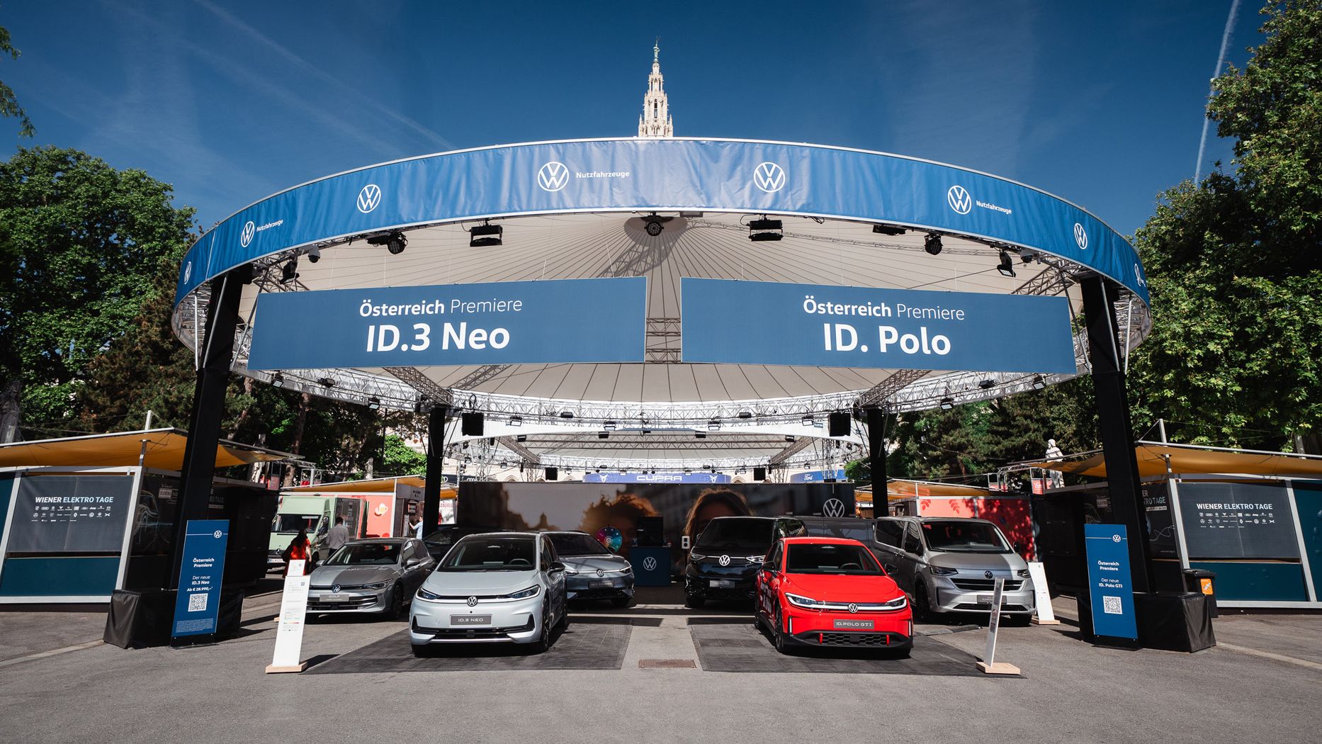 Offener VW-Ausstellungsstand im Freien mit mehreren Elektro- und Hybridfahrzeugen, im Vordergrund ein silberner, ein grauer, ein roter und ein weiterer silberner Wagen. Über dem Stand hängen große blaue Banner mit der Aufschrift „Österreich Premiere ID.3 Neo“ und „Österreich Premiere ID. Polo“. Im Hintergrund sind Bäume, Infostände und ein historisches Gebäude mit Turm zu sehen.