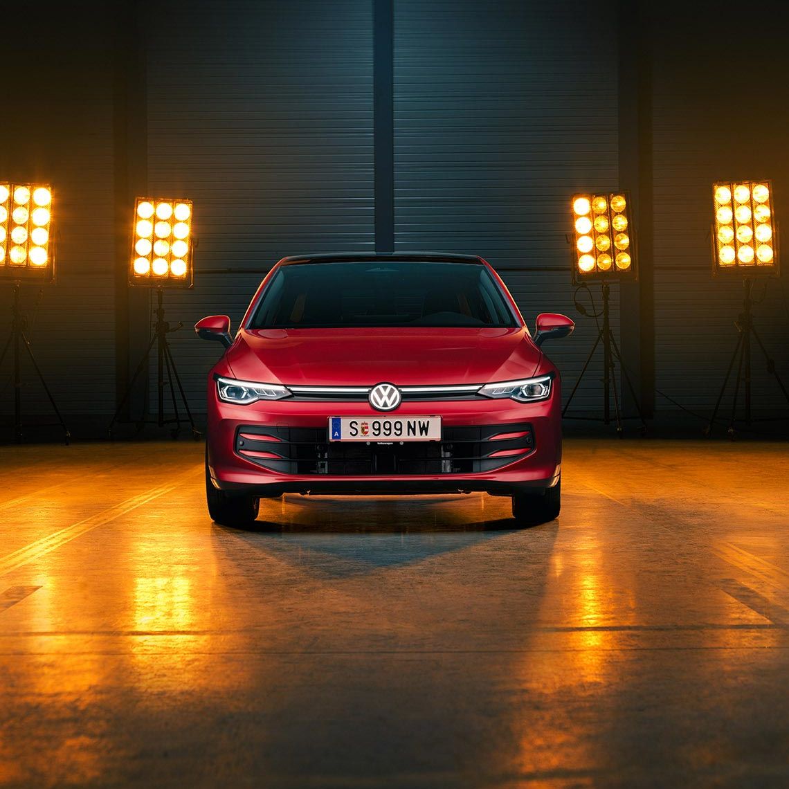 Ein roter VW Golf Falco Edition in Frontalansicht steht mittig im Bild in einer leeren Halle, Bühnenlichter strahlen das Fahrzeug von hinten an.