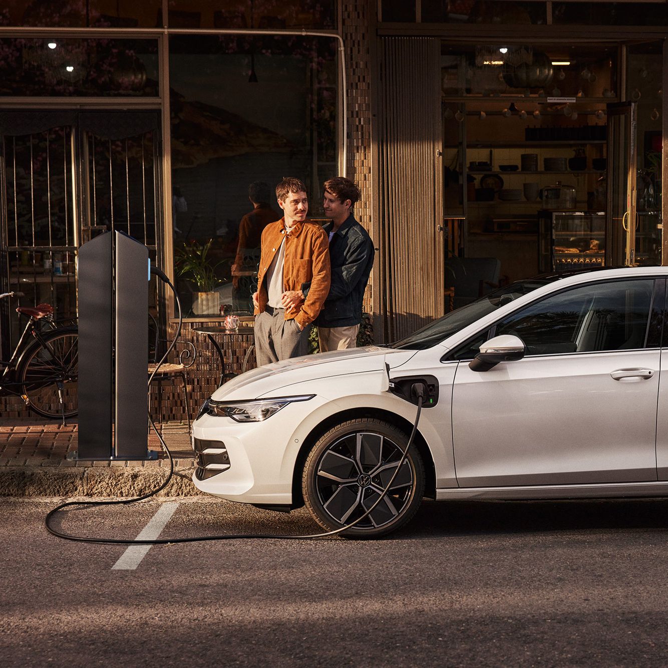 Ein weißer Golf eHybrid steht am Straßenrand und wird aufgeladen. Zwei Männer stehen neben dem Auto. 