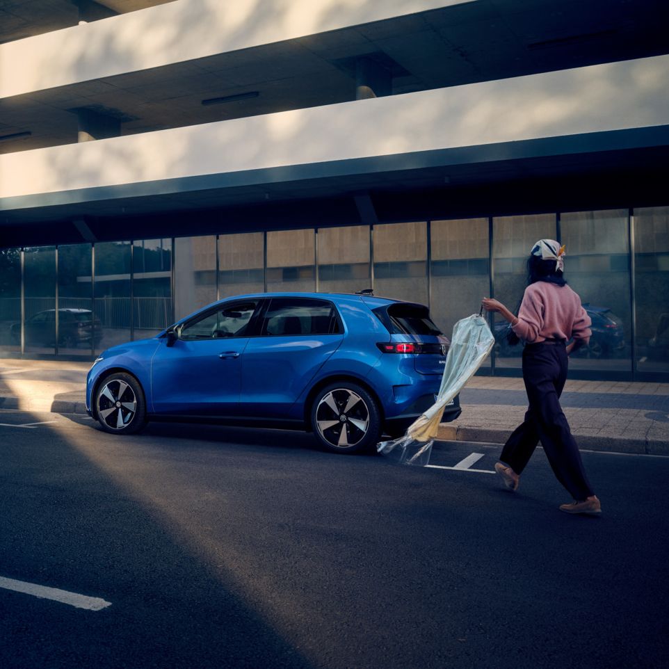 Ein blauer VW ID. Polo Style steht seitlich parkend am Straßenrand, eine Frau geht auf das Fahrzeug zu.
