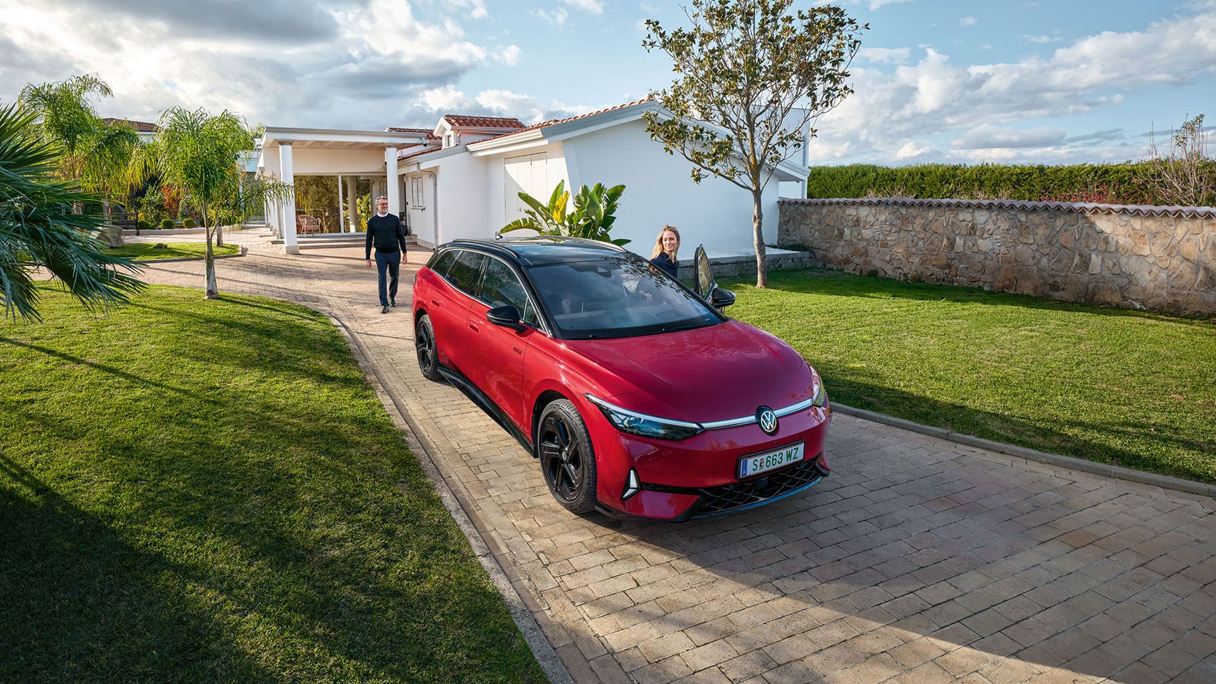 Ein roter VW ID.7 GTX parkt vor einer mediterranen Villa mit großem Garten, eine Frau steigt ein.