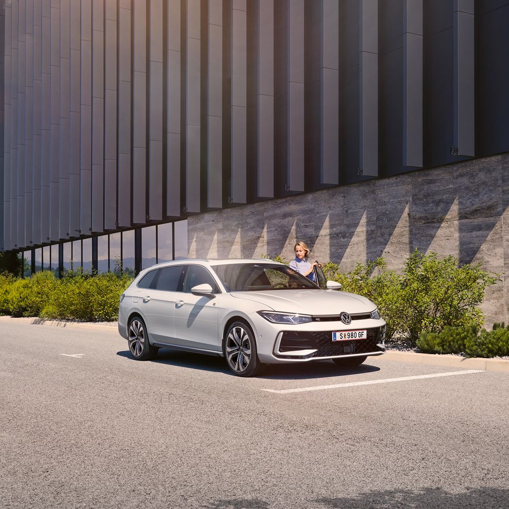 Eine Frau öffnet die Fahrertür eines weißen VW Passat R-Line und steigt ein. Im Hintergrund sieht man ein Gebäude.