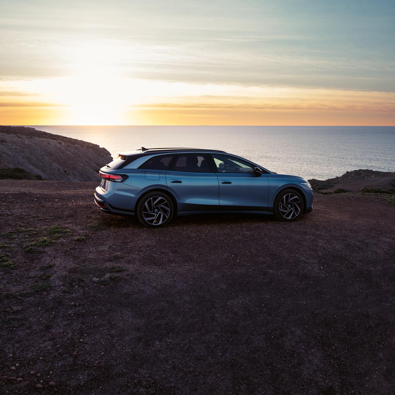 Ein blauer VW ID.7 Tourer steht oben auf einer Klippe im Sonnenuntergang, dahinter das Meer.