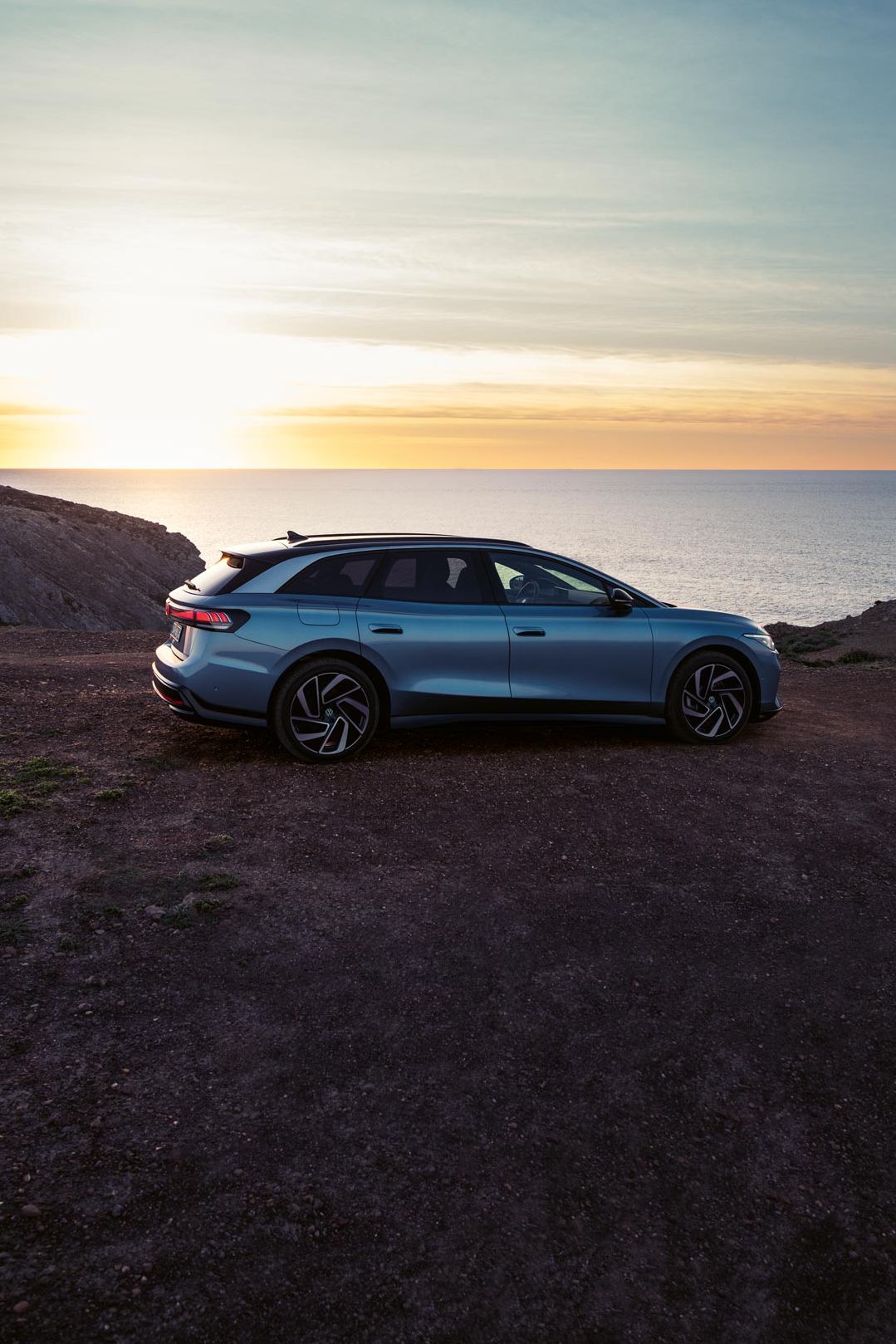 Ein blauer VW ID.7 Tourer steht oben auf einer Klippe im Sonnenuntergang, dahinter das Meer.