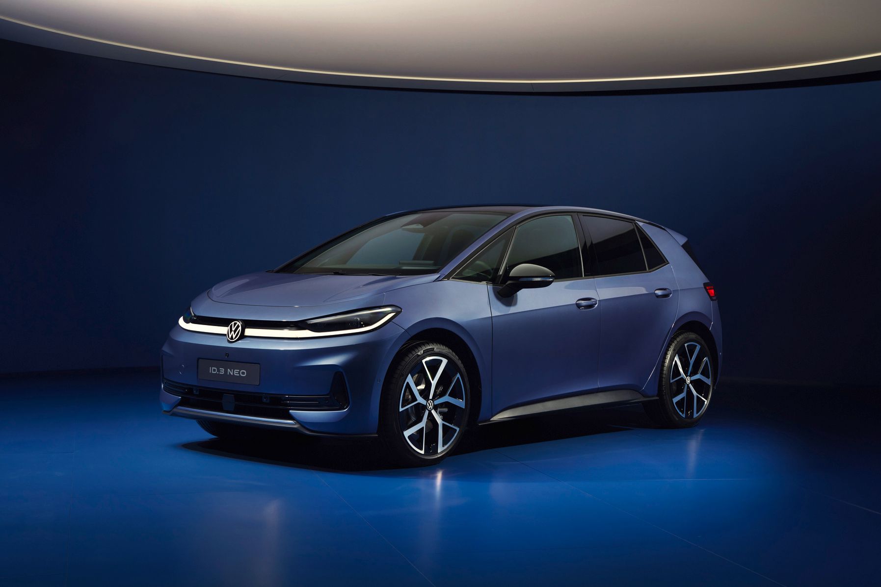 Ein dunkelblauer VW ID.3 NEO steht in einem futuristisch beleuchteten, komplett blauen Studio. Das Elektroauto ist schräg von vorne fotografiert, sodass Front und Fahrerseite gut sichtbar sind. Die Szene wirkt minimalistisch und hochwertig, mit weichem Licht und spiegelndem Boden.