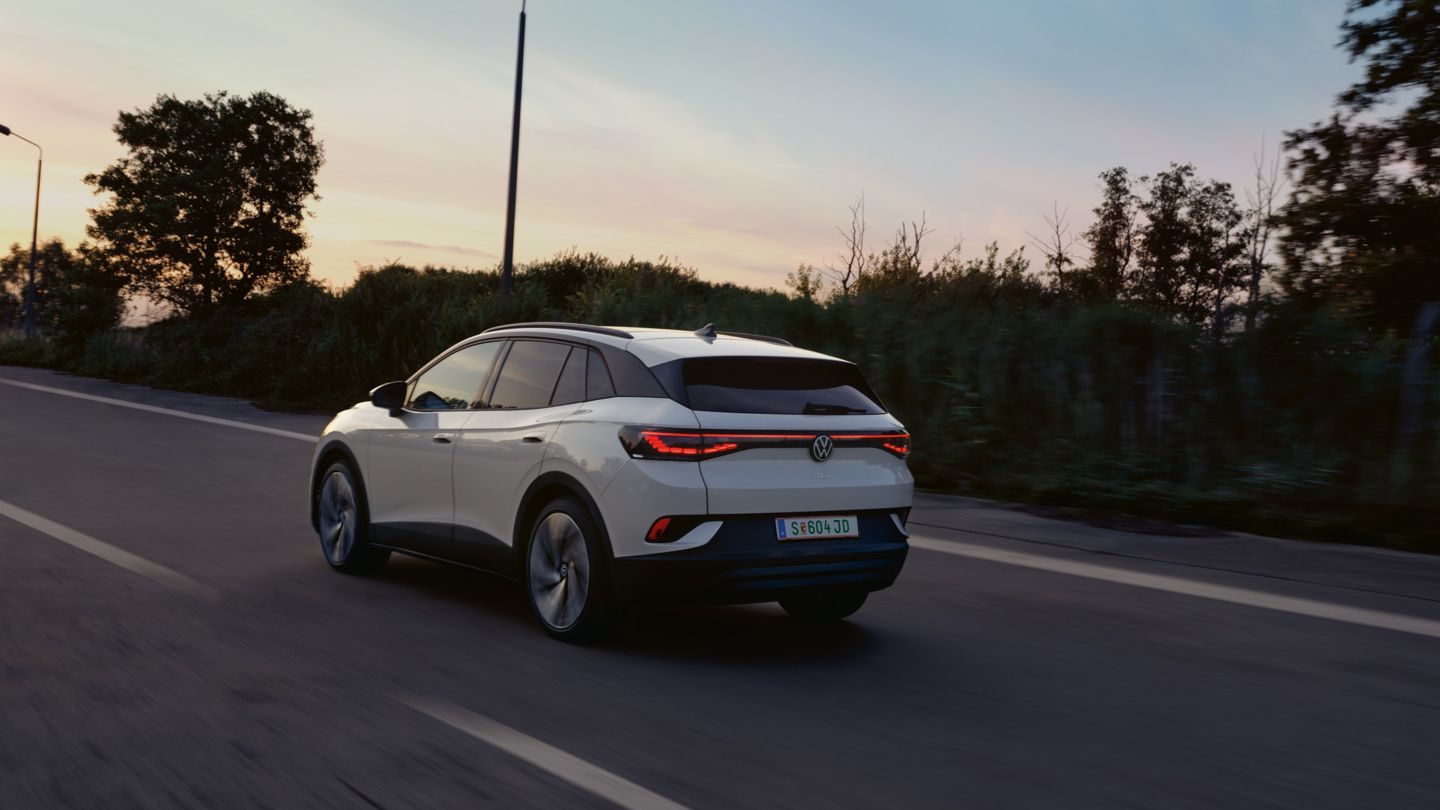 Ein weißer VW ID.4 fährt auf einer Landstraße bei Sonnenuntergang. Das Auto ist von hinten zu sehen. Die Straße ist von Bäumen und Sträuchern gesäumt.