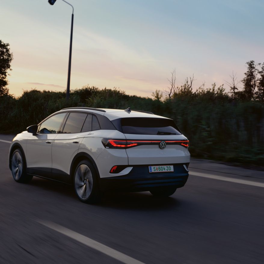 Ein weißer VW ID.4 fährt auf einer Landstraße bei Sonnenuntergang. Das Auto ist von hinten zu sehen. Die Straße ist von Bäumen und Sträuchern gesäumt.