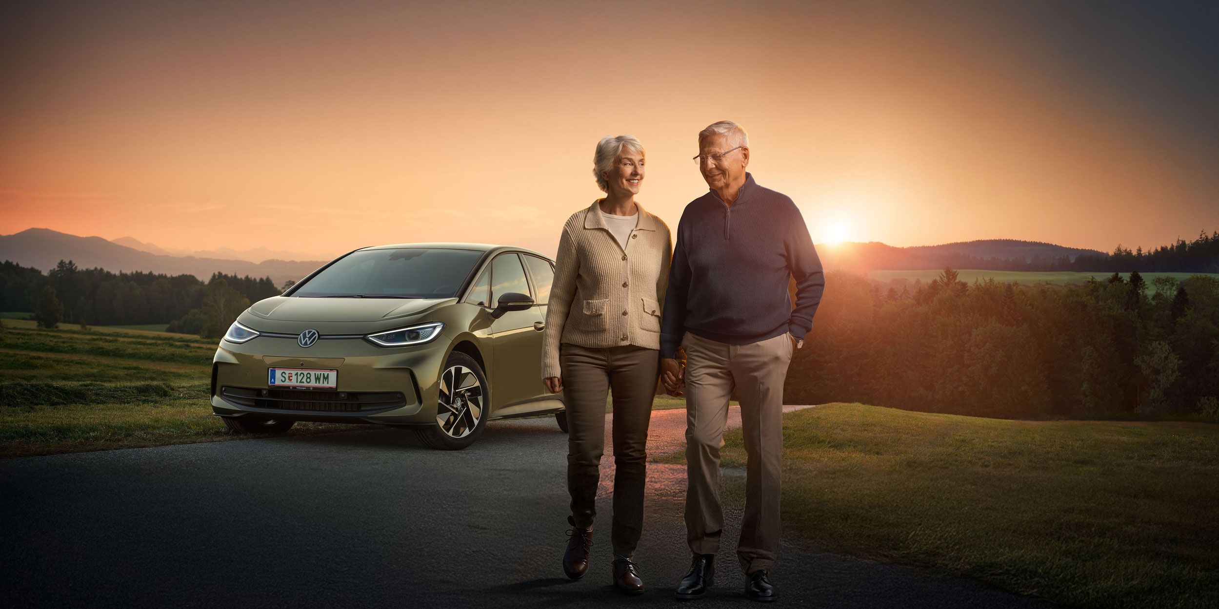 Ein älteres Paar spaziert zusammen auf einer Landstraße bei Sonnenuntergang. Im Hintergrund steht ein Auto, ein Volkswagen ID.3. Die Landschaft ist grün und hügelig.
