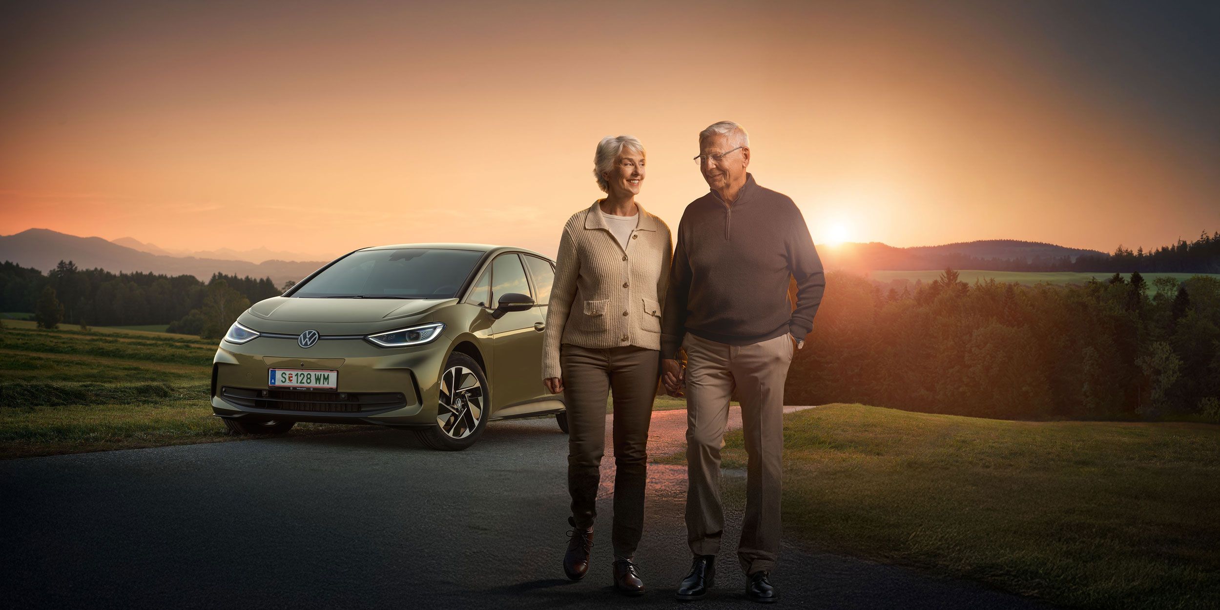 Ein älteres Paar spaziert zusammen auf einer Landstraße bei Sonnenuntergang. Im Hintergrund steht ein Auto, ein Volkswagen ID.3. Die Landschaft ist grün und hügelig.