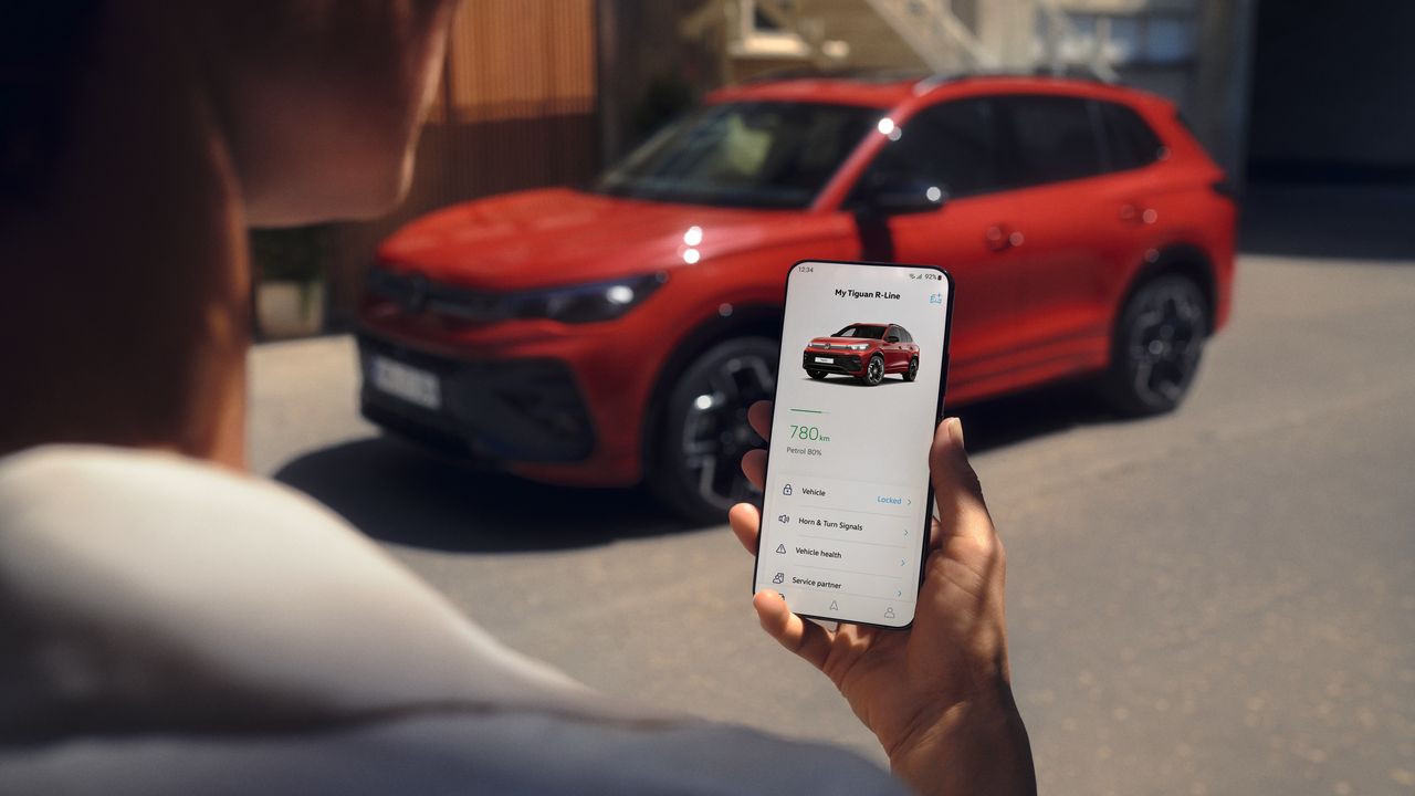 Blick über die Schulter einer Person auf ein Handy, auf dem die Volkswagen App geöffnet ist. Im Hintergrund steht ein roter Tiguan R-Line.