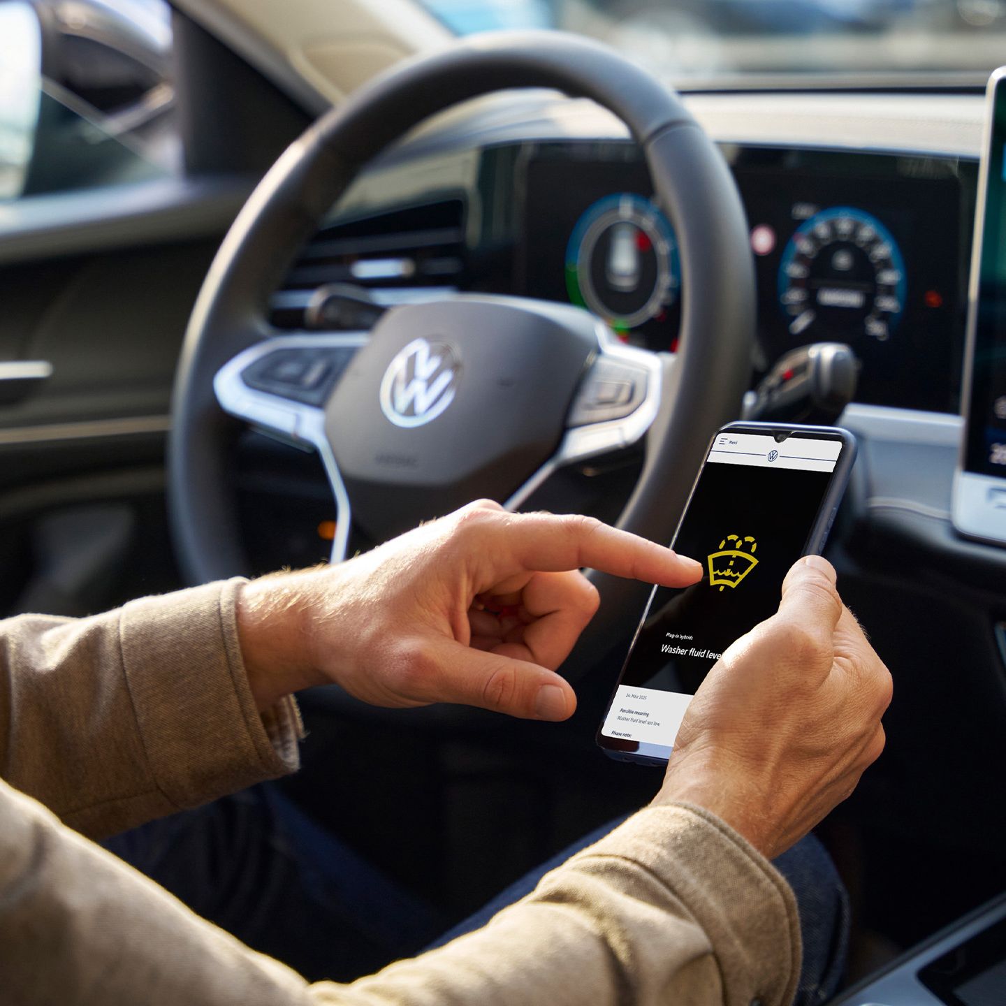 Eine Person sitzt in einem Auto und bedient ein Smartphone. Im Hintergrund ist das Lenkrad und das Armaturenbrett mit einem Bildschirm zu sehen.