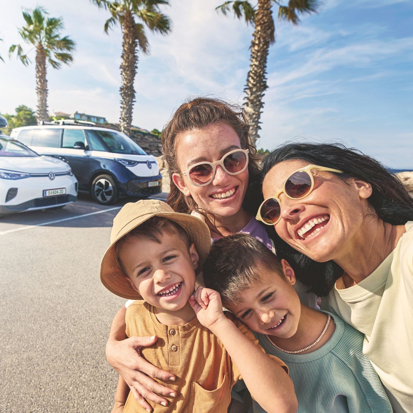 Eine Familie macht ein Selfie auf einem sonnigen Parkplatz mit Palmen im Hintergrund. Im Hintergrund stehen zwei geparkte Volkswagen mit Gepäck auf dem Dach.