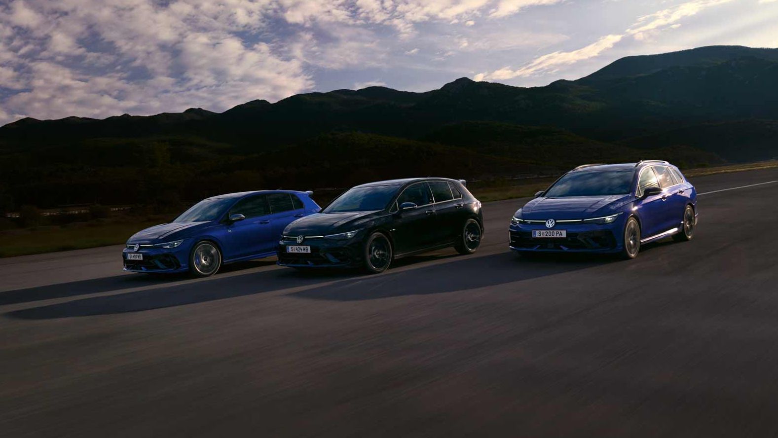 Drei VW Golf R fahren nebeneinander auf einer Straße bei Sonnenuntergang. Die Himmel zeigt einige Wolken und im Hintergrund sind Berge sichtbar. Die Farben der Autos sind blau, schwarz und dunkelblau.