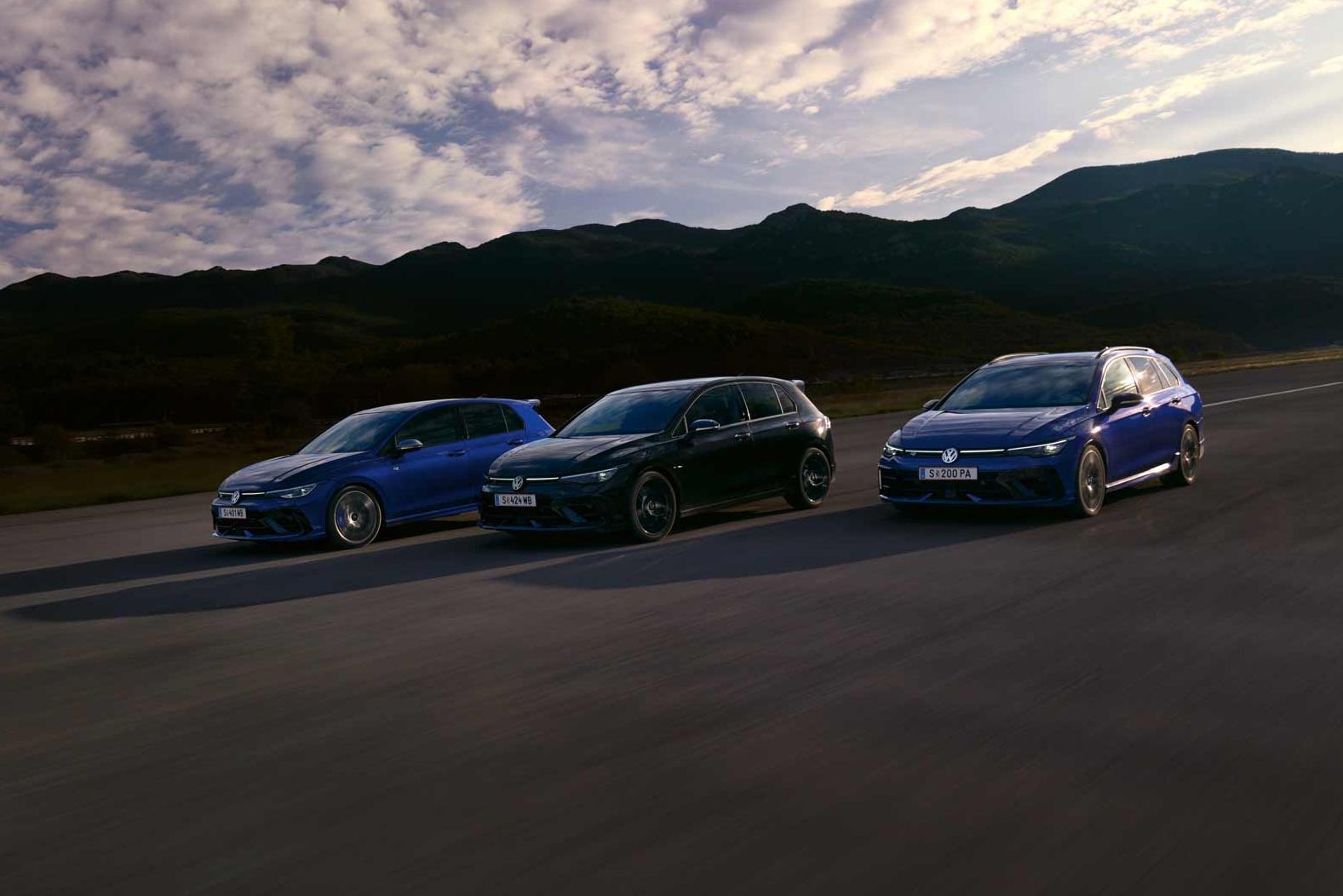 Drei VW Golf R fahren nebeneinander auf einer Straße bei Sonnenuntergang. Die Himmel zeigt einige Wolken und im Hintergrund sind Berge sichtbar. Die Farben der Autos sind blau, schwarz und dunkelblau.