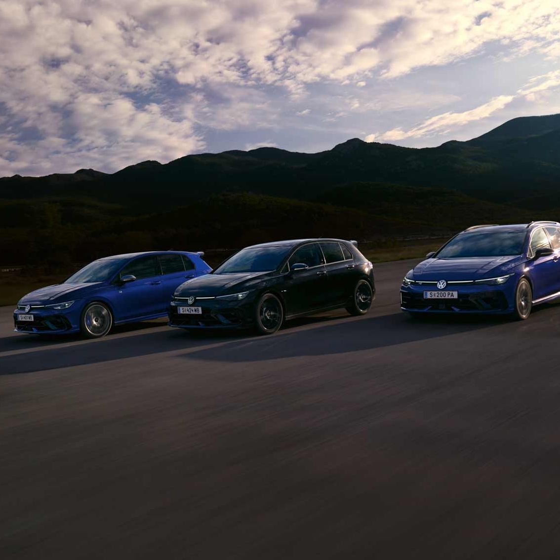 Drei VW Golf R fahren nebeneinander auf einer Straße bei Sonnenuntergang. Die Himmel zeigt einige Wolken und im Hintergrund sind Berge sichtbar. Die Farben der Autos sind blau, schwarz und dunkelblau.