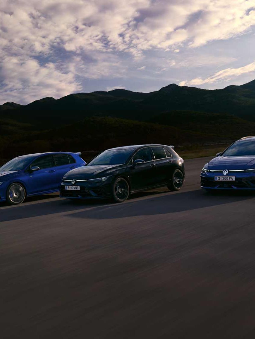 Drei VW Golf R fahren nebeneinander auf einer Straße bei Sonnenuntergang. Die Himmel zeigt einige Wolken und im Hintergrund sind Berge sichtbar. Die Farben der Autos sind blau, schwarz und dunkelblau.