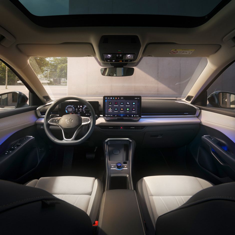 Das Bild zeigt das Innere eines T-Roc mit einem Sonnen- oder Glasdach. Das Cockpit hat ein großes Bildschirm-Display und ein Lenkrad mit mehreren Funktionen. Außen ist eine Grünfläche und ein Gebäude zu erkennen.