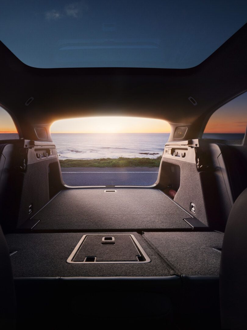 Blick aus dem geöffneten Kofferraum mit umgeklappter Rücksitzbank aus dem ID.7 Tourer auf einen Sonnenuntergang über dem Meer. 