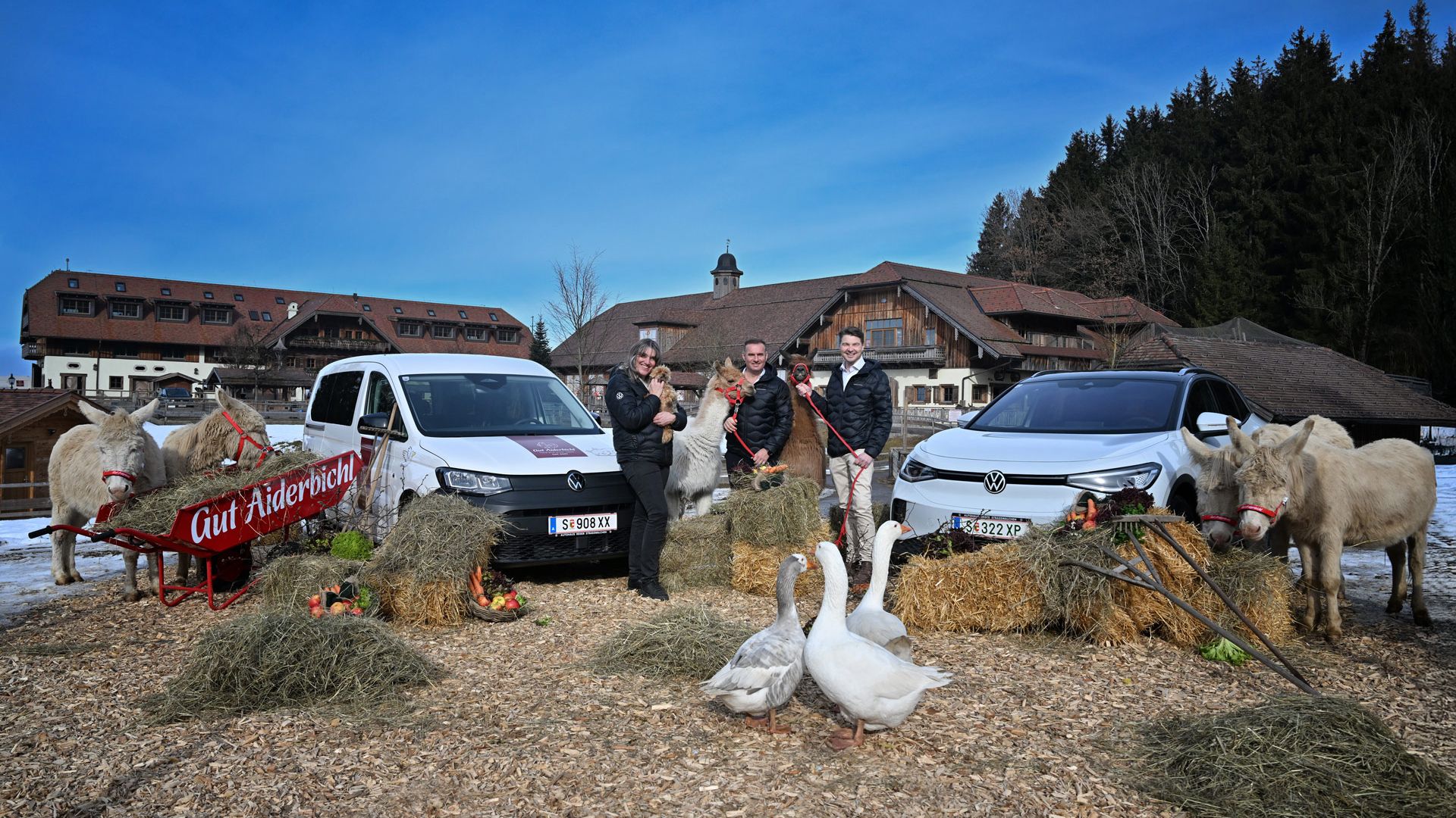 Mehrere Personen stehen in einem landwirtschaftlichen Umfeld mit Heuballen, einem Schlitten und zwei weißen Autos. Im Vordergrund sind mehrere Gänse und Pferde zu sehen. Im Hintergrund stehen mehrere Gebäude, die wie ein Bauernhof aussehen.